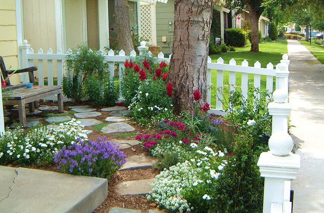 a white picket fence next to a tree and flower bed in front of a house