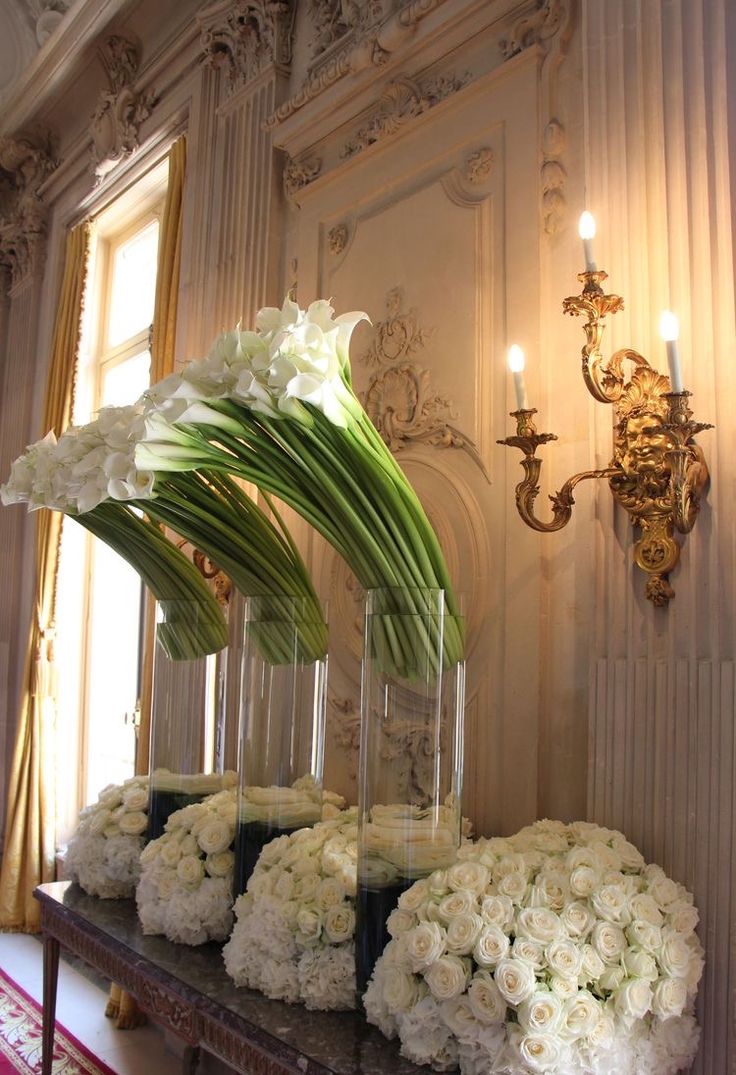 white flowers are in vases on a table next to a chandelier and wall sconces