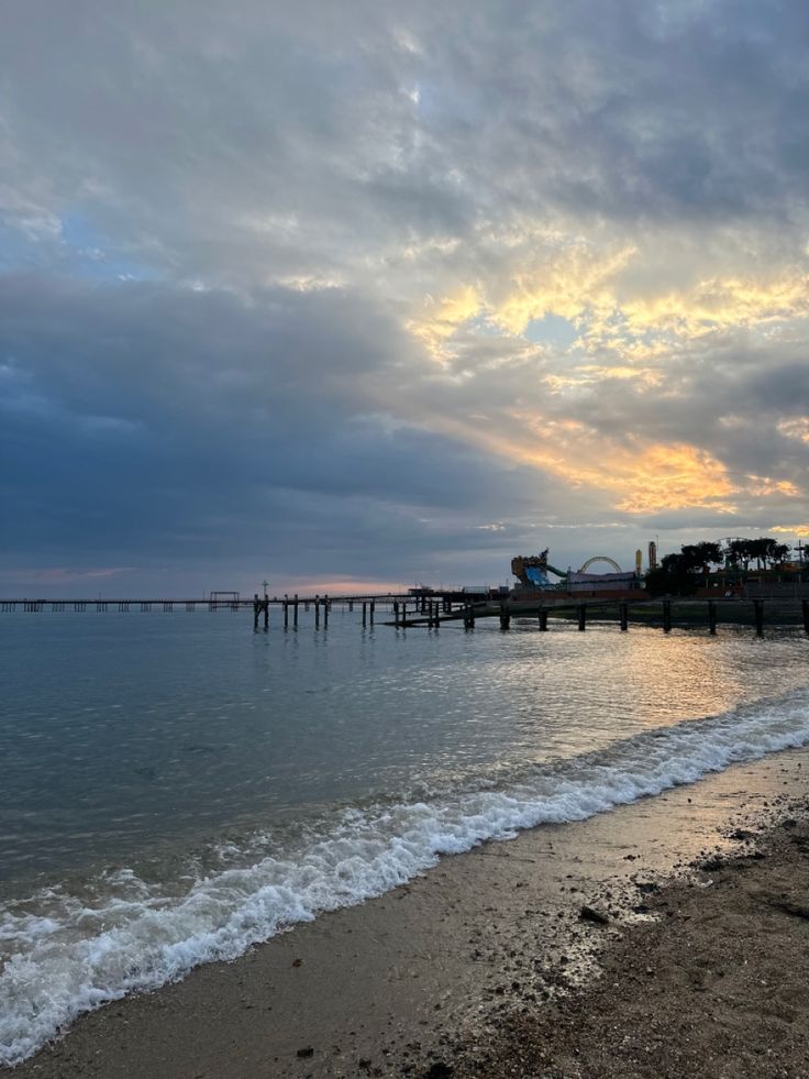 the sun is setting over the water at the beach with a pier in the distance