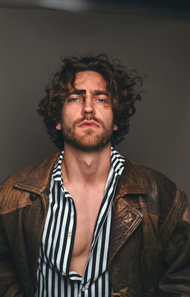 a man with long curly hair wearing a brown jacket and striped shirt is looking at the camera