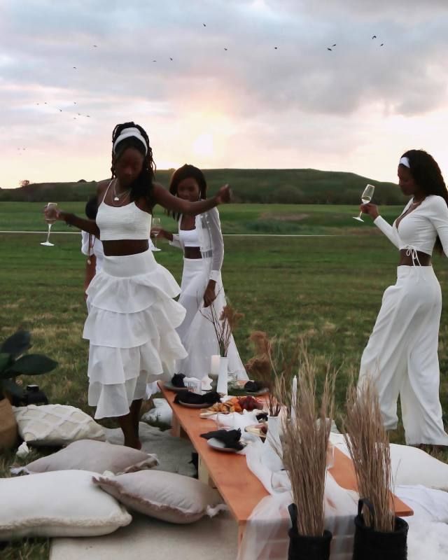 three women dressed in white dancing around a table with food and wine glasses on it