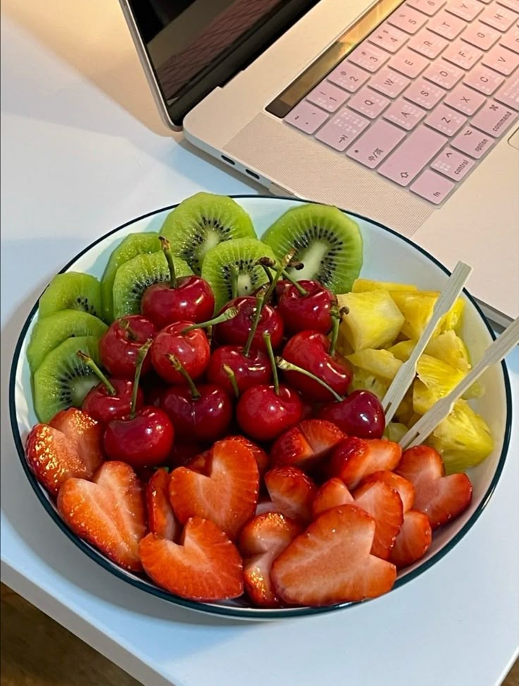 a bowl of fruit with strawberries, kiwis and cherries in front of a laptop