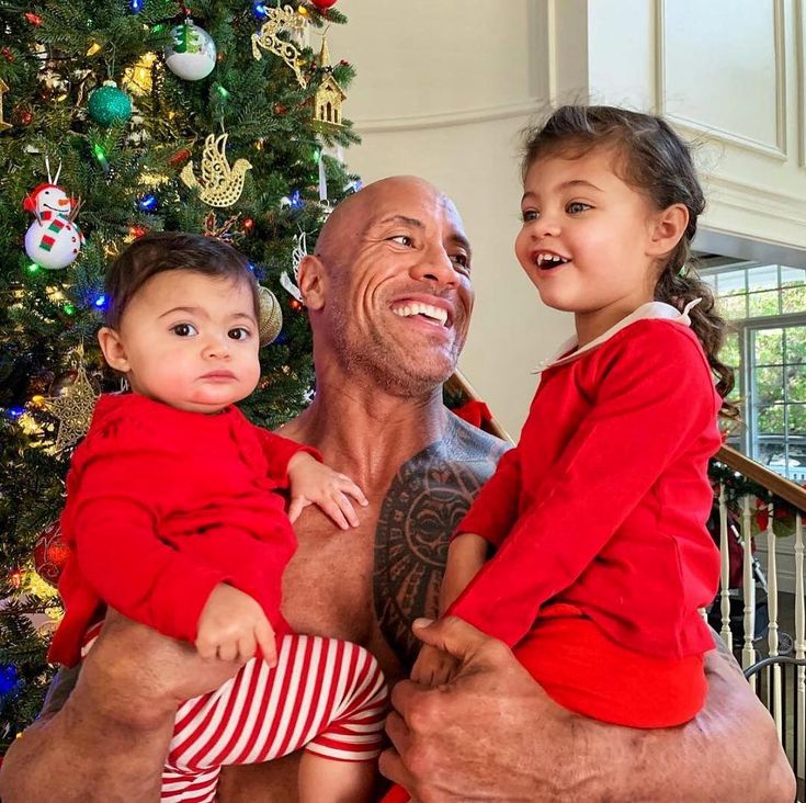 a bald man holding two children in front of a christmas tree with decorations on it