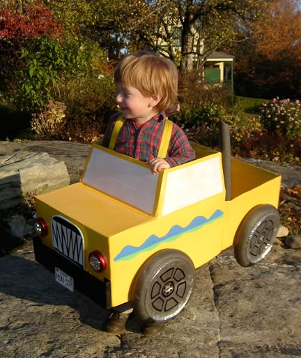 a young boy riding in a yellow toy truck on top of a rocky surface with trees and bushes behind him