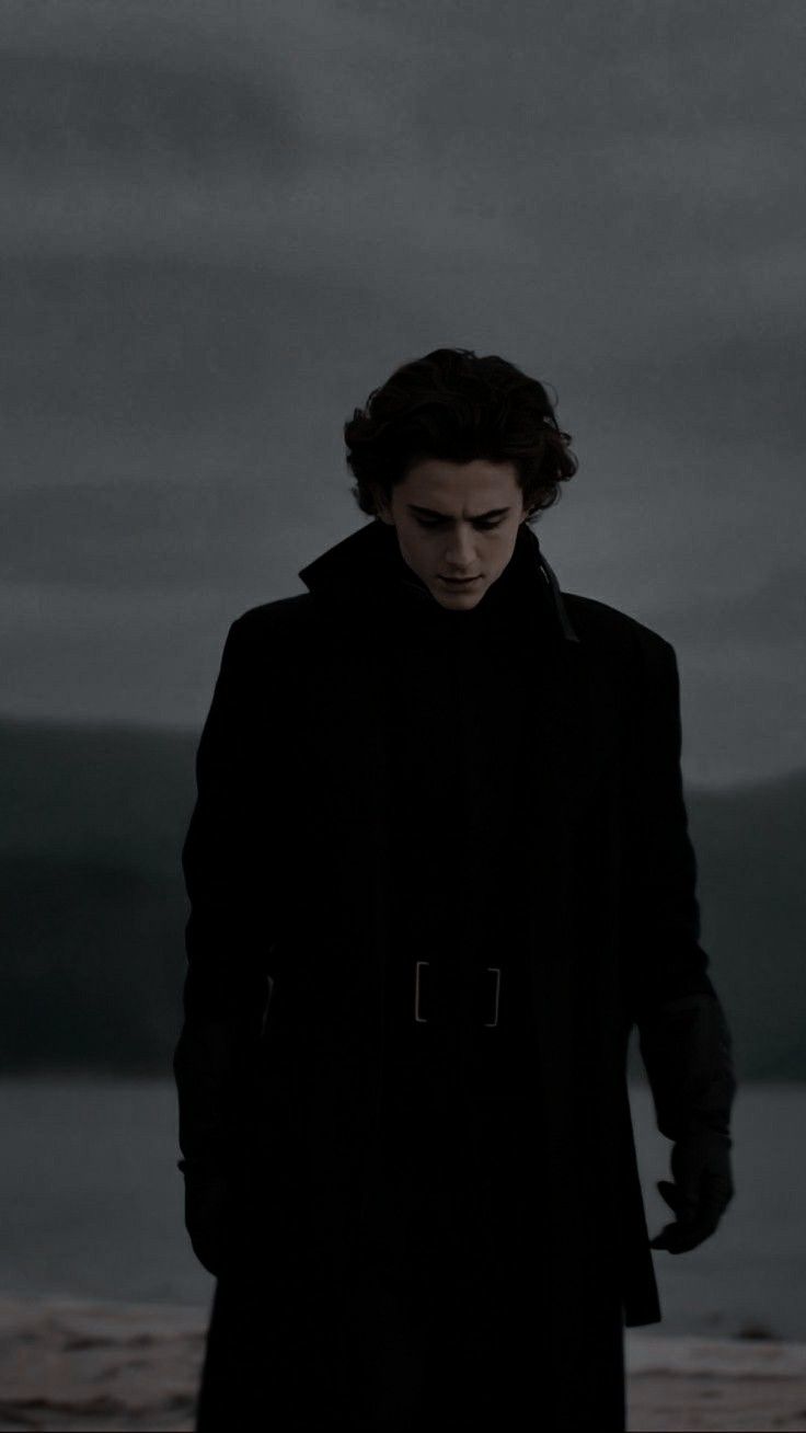 a man standing on top of a beach next to the ocean in a black coat