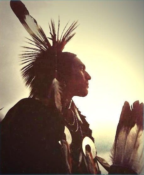 a native american man standing in front of the sun with his headdress on