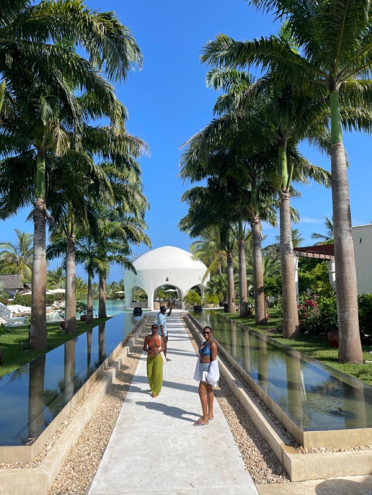 two women walking down a walkway between palm trees and the water in front of them