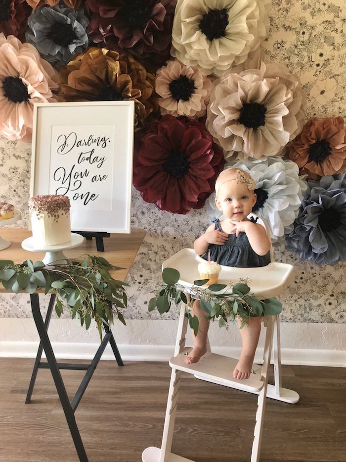 a baby sitting in a high chair with flowers on the wall behind it