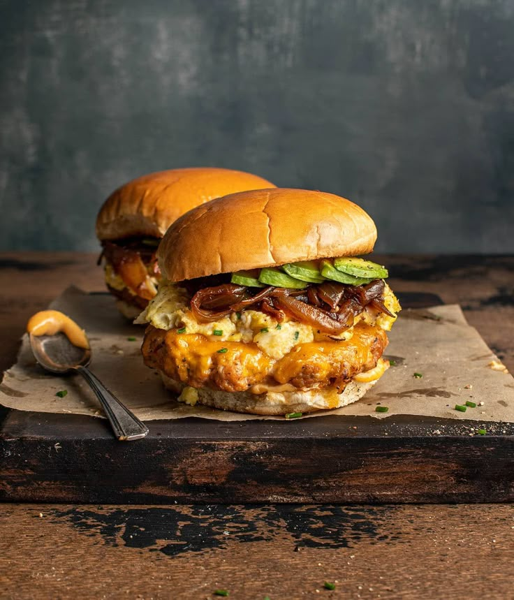 two burgers sitting on top of a cutting board next to a spoon and fork