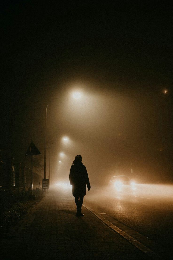 a person walking down a street at night in the fog with cars driving on it