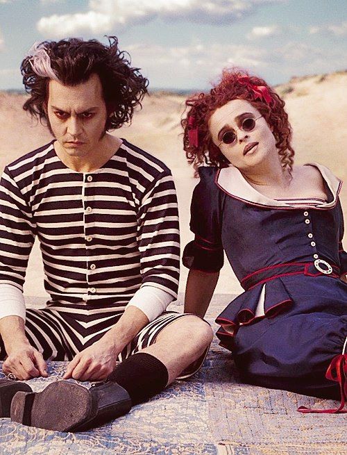 two people sitting on the ground in front of sand dunes, one is wearing sunglasses