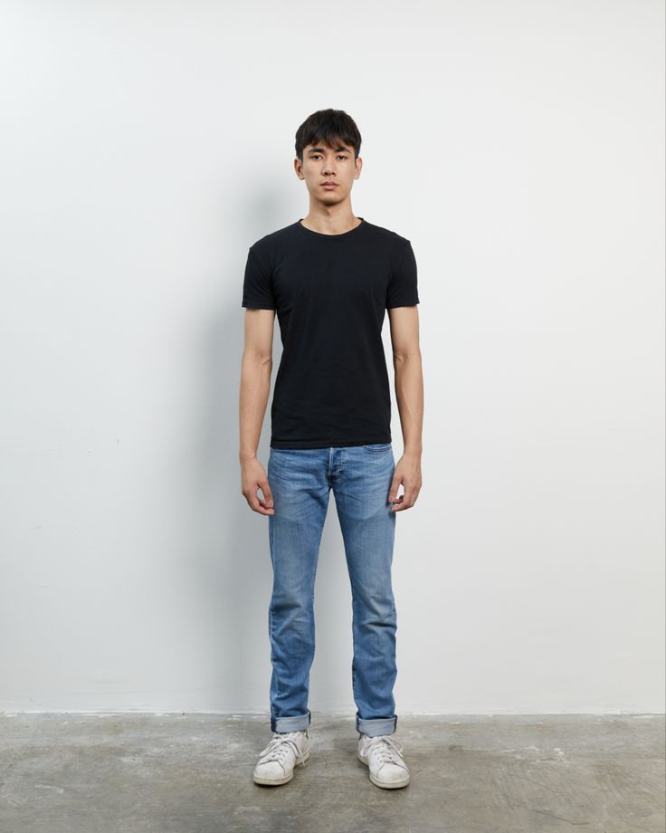 a man standing in front of a white wall wearing jeans and a black t - shirt