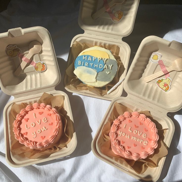 four small birthday cakes in plastic containers on a white sheet covered table with the words happy birthday written on them