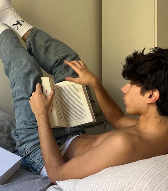 a man laying in bed reading a book with his feet up on the pillow while holding an open book