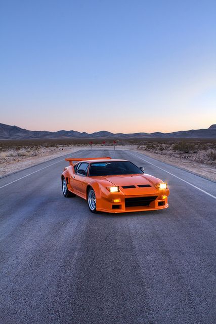 an orange sports car is driving down the road in the middle of the desert at sunset