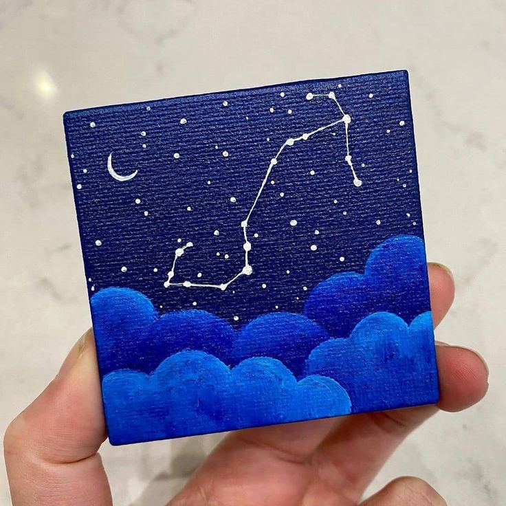 a hand holding up a small square painted with the zodiac sign on it's side