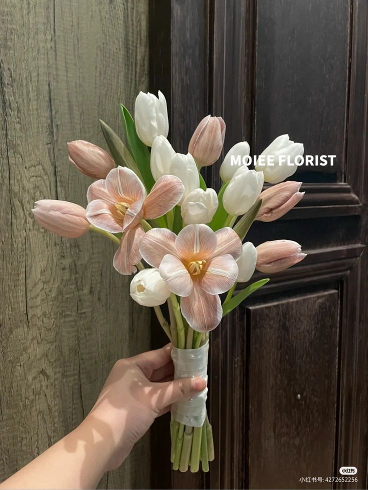 a hand holding a bouquet of white and pink tulips in front of a wooden door