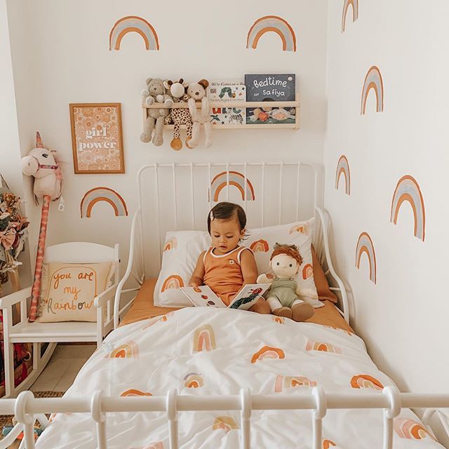 a small child sitting on a bed with two stuffed animals in front of him and the wall has rainbows painted on it