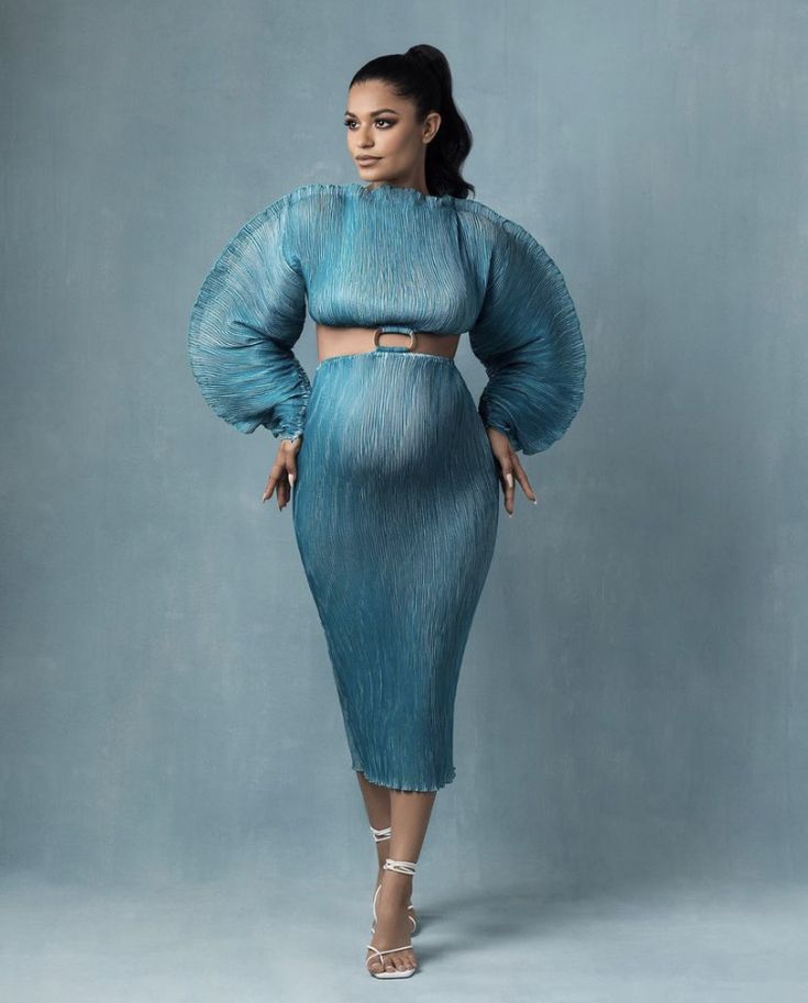 a woman in a blue dress and heels standing on a gray background with her hands on her hips