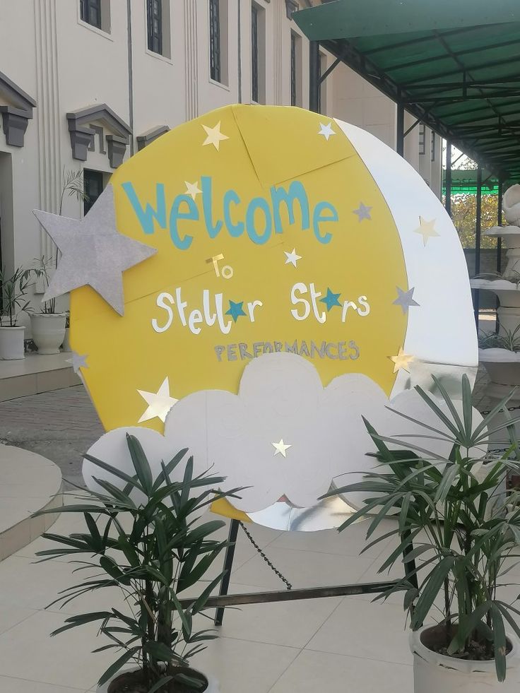 a welcome sign in front of a building with potted plants on the ground and a large yellow moon behind it