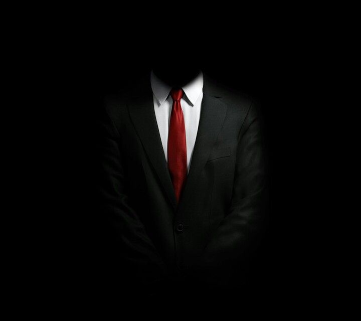 a man in a suit and red tie standing in the dark with his arms crossed