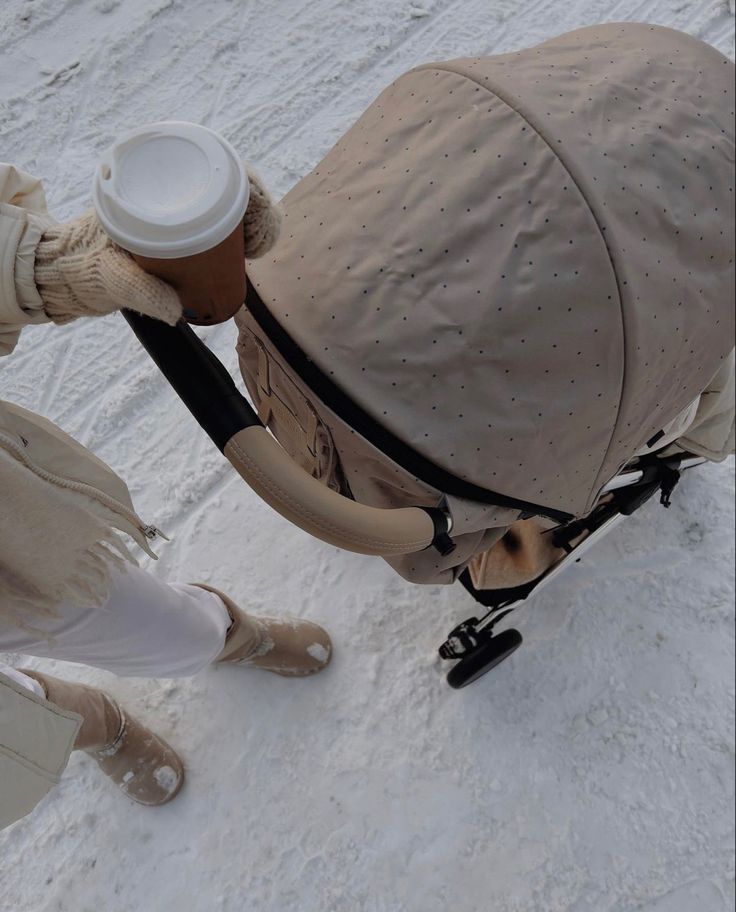 a woman holding a coffee cup in her right hand and pushing a stroller on the snow