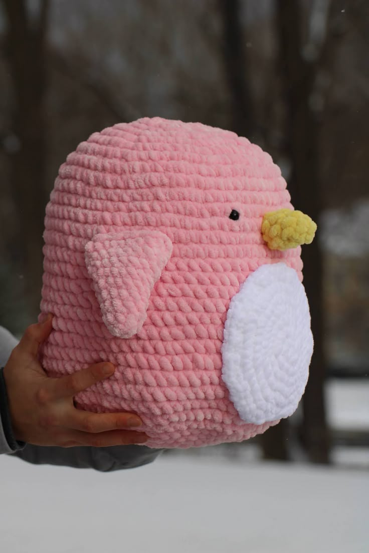 a hand holding a pink crocheted stuffed animal in front of snow covered trees