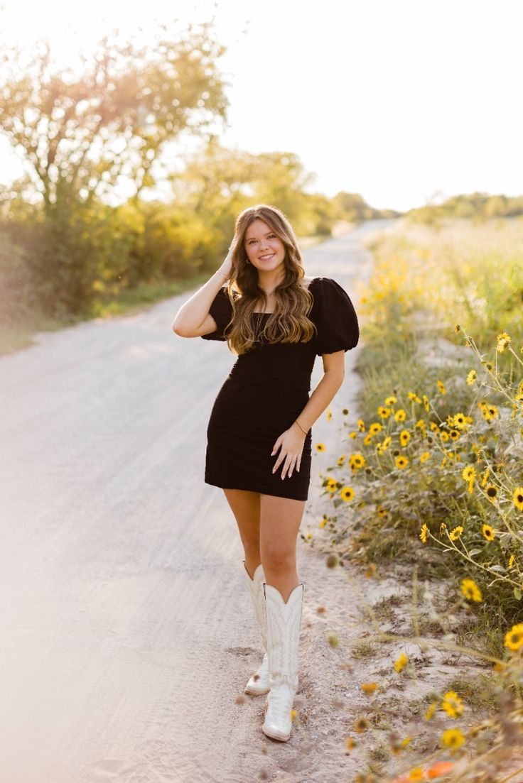 a woman standing in the middle of a dirt road with sunflowers behind her