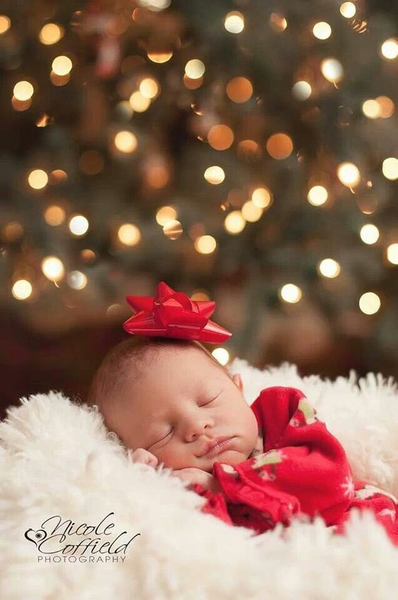 a newborn baby wearing a red bow laying in front of a christmas tree