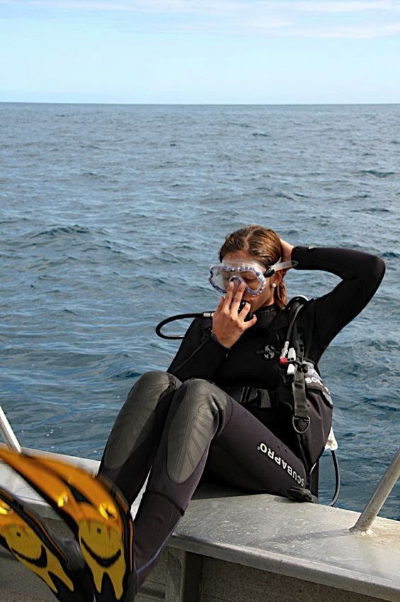 a woman in scuba gear sitting on the edge of a boat talking on her cell phone