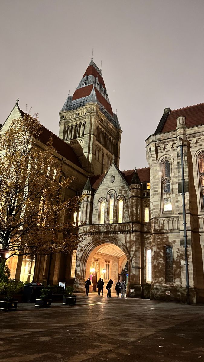 people are walking in front of an old building at night with the lights on and there is a clock tower