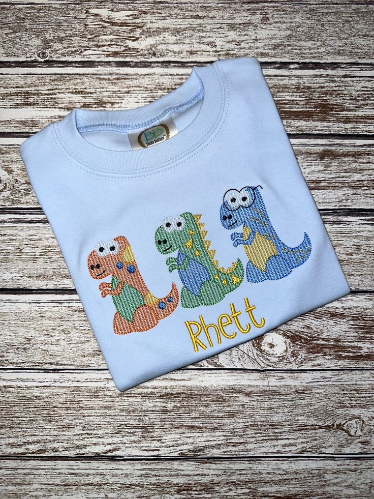 Boy's light blue short or long sleeve sleeve shirt with embroidered dinos!  Name is stitched in yellow.    The shirts are thicker than the average t'shirt and you will not be disappointed in the quality. I use shirts from 2 different blanks companies who offer quality shirts.  Unless you have a preference, I will use whichever I have in stock in the size ordered. All shirts in my shop are finished on the inside with a soft covering to protect the skin against the underside of the design.   All items listed in my shop are made with care and in a pet free, smoke free shop. Thank you so much for shopping with Jus4boys and if you have any questions, please ask.  I respond quickly unless it is late night. Blue Dinosaur Print Long Sleeve Top, Blue Long Sleeve Dinosaur Print Top, Fun Long Sleeve Shirt For Summer, Long Sleeve Blue Top With Dinosaur Print, Blue Long Sleeve Top With Dinosaur Print, Fun Long Sleeve Summer Shirt, Fun Summer T-shirt With Dinosaur Print, Summer Cotton Shirt For Birthday, Cotton Shirt For Birthday In Summer