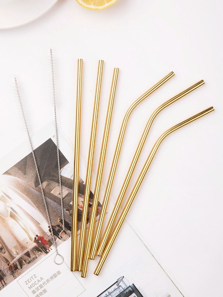 four gold straws sitting on top of a white table next to a plate with an orange slice