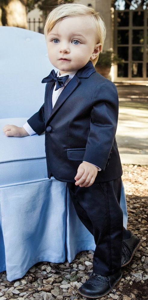 a little boy in a suit and bow tie standing next to a blue chair outside