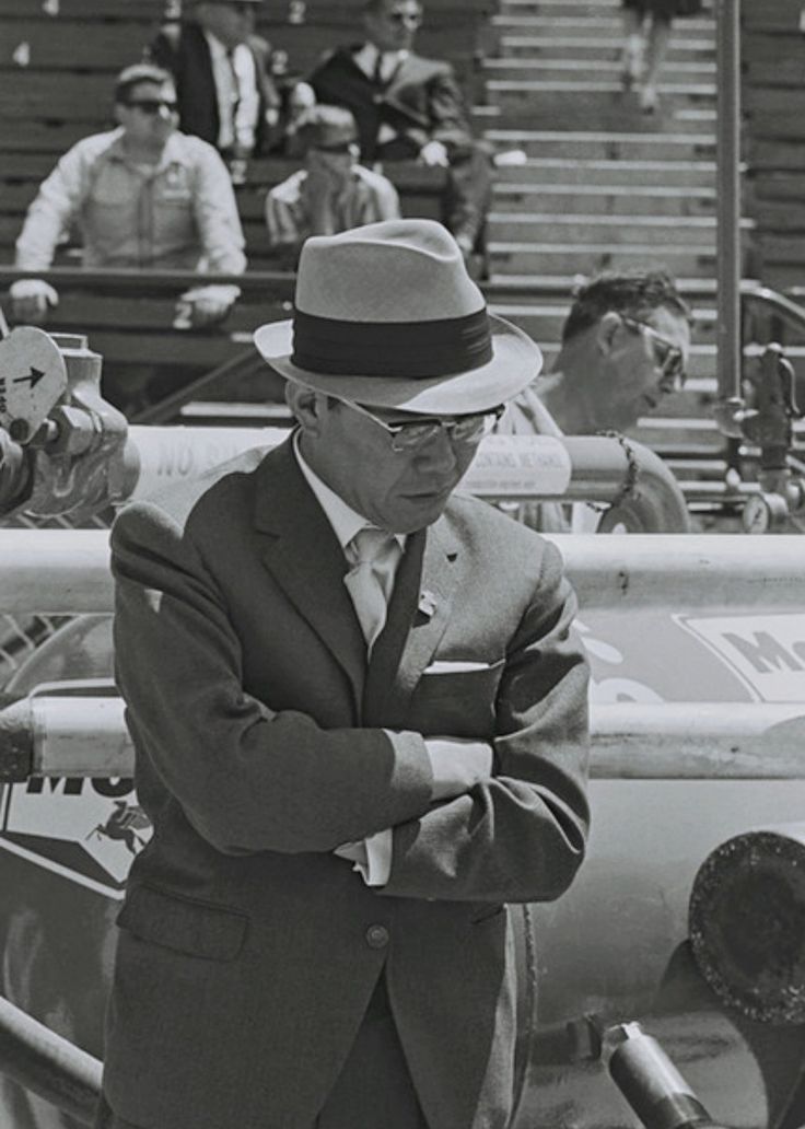 a man in a suit and hat standing next to a boat with people sitting on the bleachers