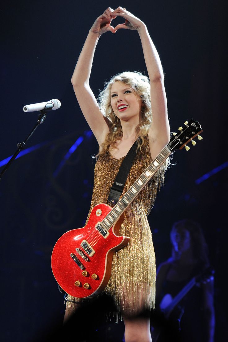 a woman in a gold dress holding a red guitar and making the v sign with her hands