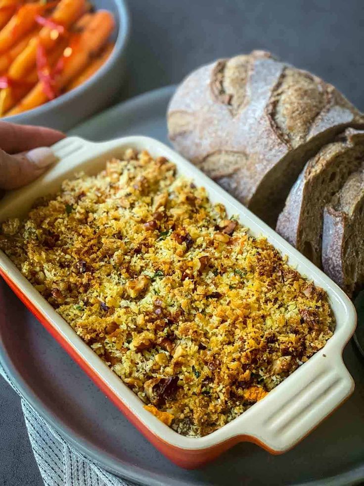 a person holding a spoon over a casserole dish with carrots and bread in the background