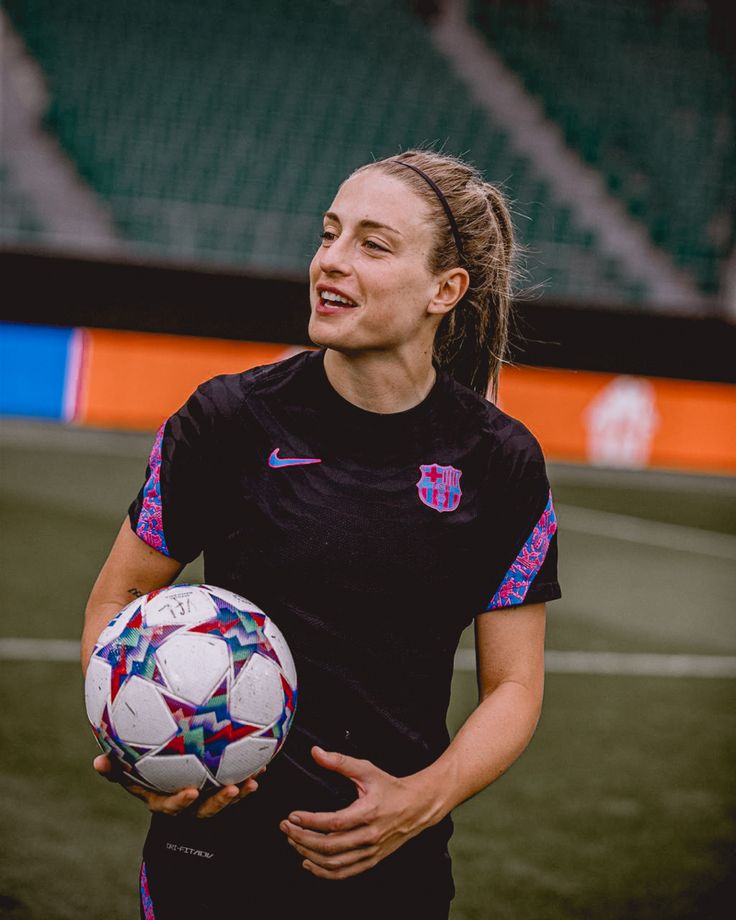 a woman holding a soccer ball on top of a grass covered field in front of a stadium