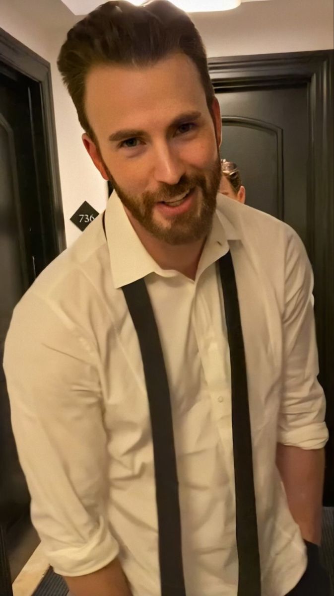 a man wearing a white shirt and black tie standing in front of a door with his hands on his hips