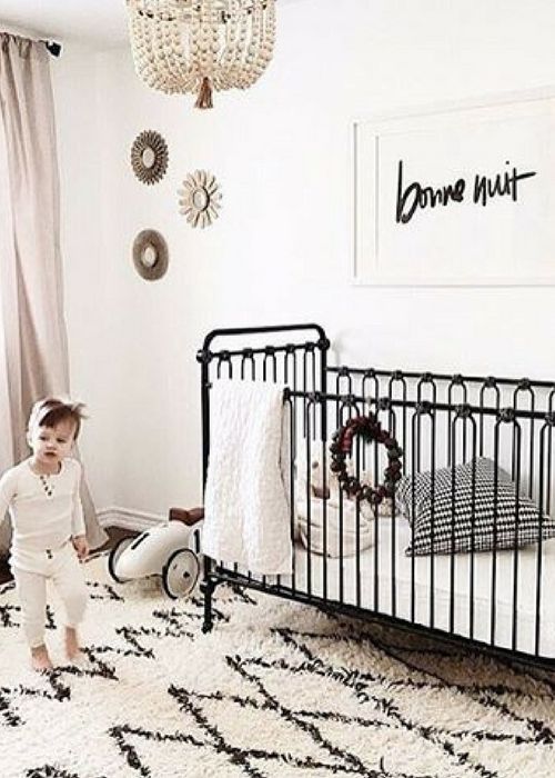 a baby standing next to a black crib in a white room with a chandelier hanging from the ceiling