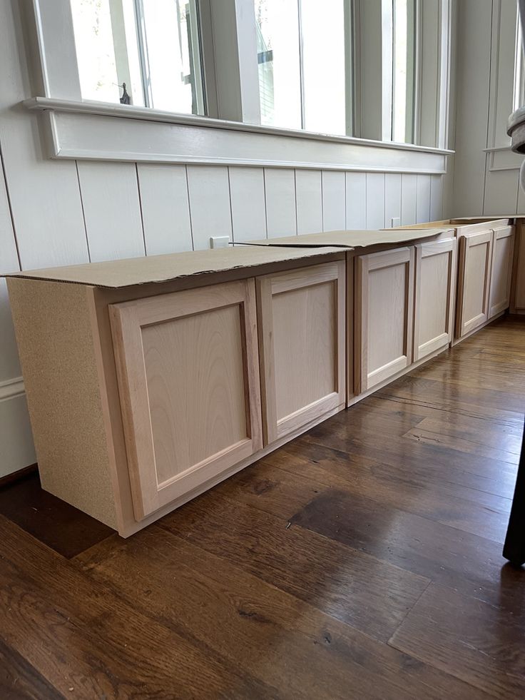 an empty kitchen with wooden floors and white cabinets on the wall, in front of a window