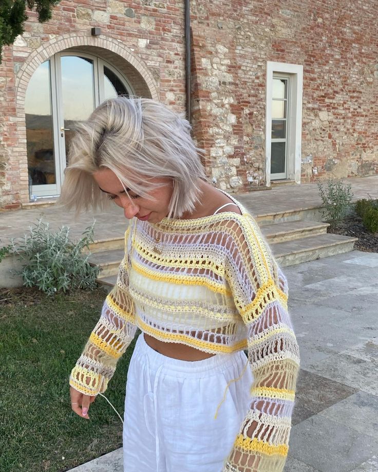 a woman with blonde hair wearing a yellow and white crochet sweater standing in front of a brick building