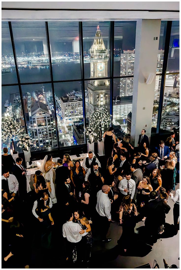 a large group of people standing in front of a window at night with the city lights lit up