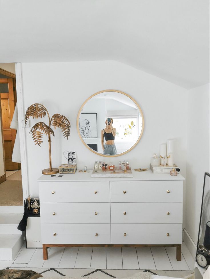 a woman standing in front of a mirror on top of a dresser next to a plant