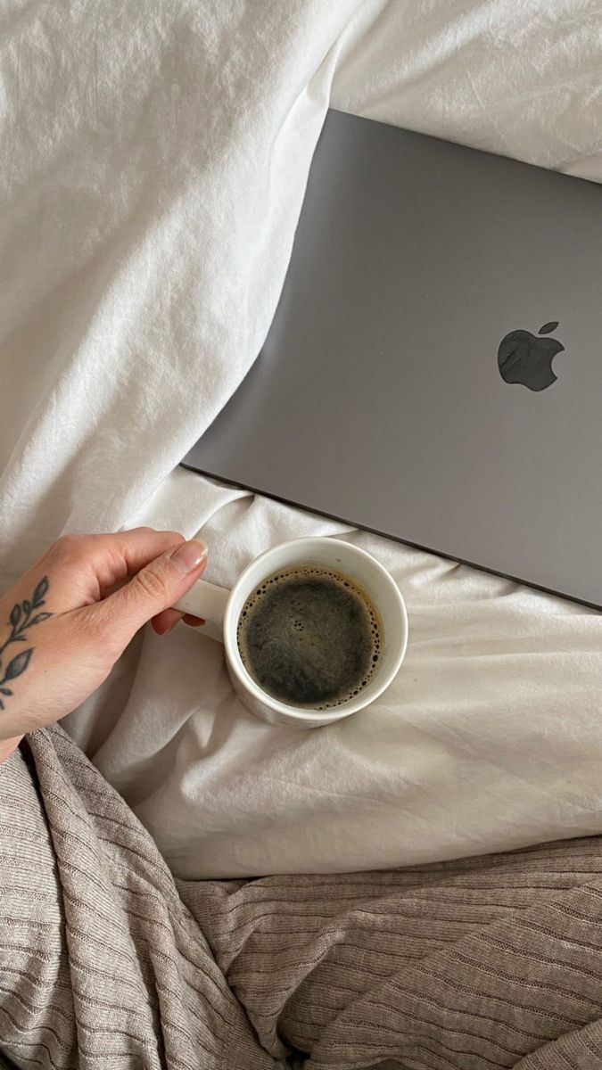 a person with their hand on a coffee cup next to an apple laptop in bed