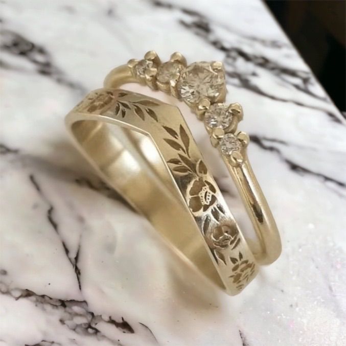 two gold wedding rings sitting on top of a marble counter with flowers and leaves in the middle