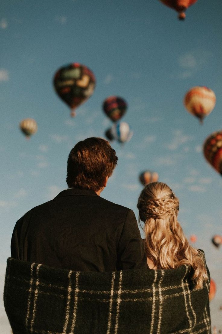 a man and woman sitting on top of a blanket looking at hot air balloons