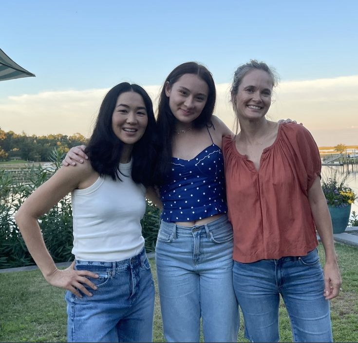 three young women standing next to each other in front of a body of water at sunset