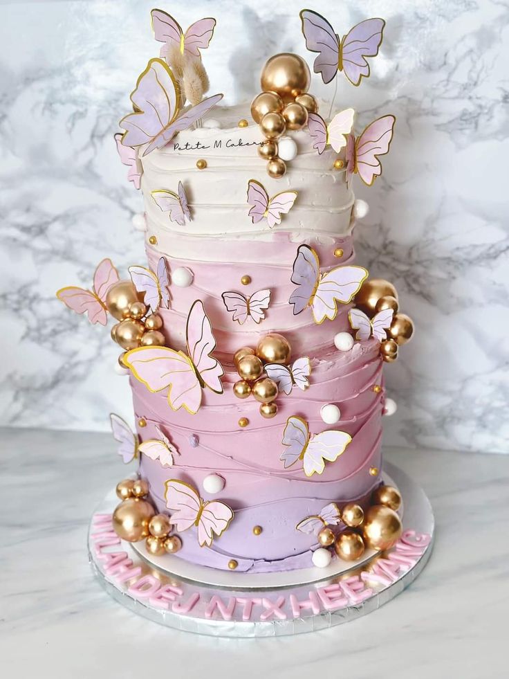 a three tiered cake with pink and gold butterflies on the top, sitting on a marble surface