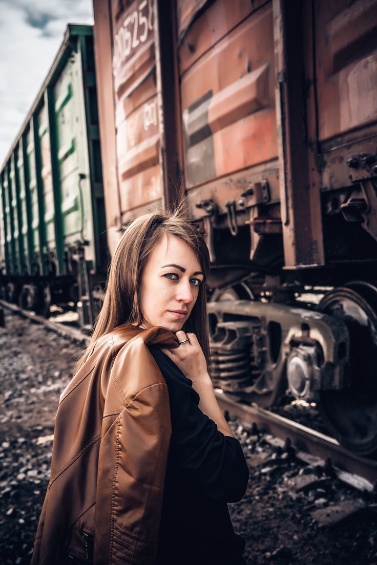 a beautiful young woman standing next to a train on the tracks with her jacket over her shoulder